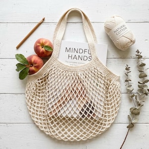 以下が含まれることがあります： 「MINDFUL HANDS」というタイトルの本が入った、クリーム色のクロシェ編みマーケットバッグ。バッグには、緑の葉がついた2つの桃、木製のクロシェフック、ナチュラルコットンの毛糸玉が添えられています。