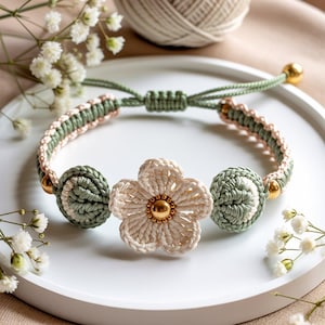 May include: A handmade macrame bracelet featuring a cream-colored flower and two green and cream-colored spheres. The bracelet has a green cord and gold-colored accents. The bracelet is displayed on a white plate with small white flowers.