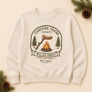 Peut inclure: Un sweat-shirt à col rond couleur crème avec un motif brodé central représentant un feu de camp et une saucisse sur un bâton. Le texte indique CAMPFIRE FRANK SOCIETY, MILLER FAMILY, PINE LAKE CABIN et EST. 2026, encadré par deux sapins.