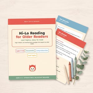 May include: A Brave Little Readers workbook titled Hi-Lo Reading for Older Readers with a red top and green bottom border, featuring a cat icon. Beside it are three coloured crayons and a small green leafy branch on a light wood surface.