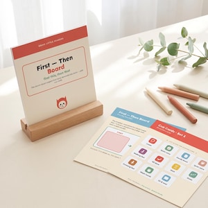 May include: A First-Then Board visual support system with a wooden stand, featuring a cream-colored card with red accents and a cat icon. Beside it lies a printed sheet of cue cards with colorful icons for tasks like listen, wash hands, and wait.