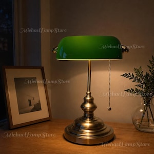 May include: A banker lamp with a polished brass base and a semi-cylindrical green glass shade. The lamp features a pull chain switch with a small spherical bead and emits a warm light. The text Michael Lamp Store is visible on the image.