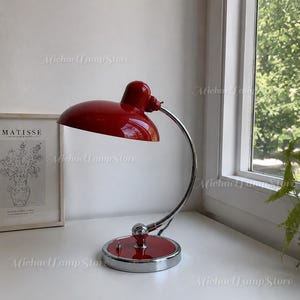 May include: A desk lamp featuring a glossy red dome-shaped shade and a matching red circular base. The lamp has a curved, polished chrome arm that connects the base to the shade, with a small toggle switch located on the top of the base.