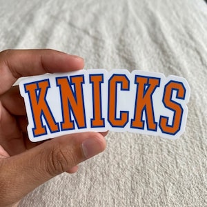 May include: A white sticker with the word "KNICKS" in orange block letters with a blue outline. The sticker is held by a hand against a neutral background.