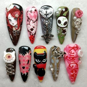 May include: A collection of ten long, pointed acrylic nails with various designs. The designs include cartoon characters, hearts, crosses, and other gothic-inspired elements. Colors include black, red, pink, white, and green. Some nails feature 3D embellishments.