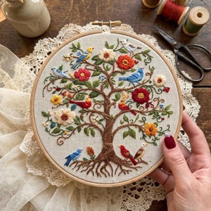 Puede incluir: Un bastidor de bordado circular de madera que contiene un panel de tela con un diseño detallado cosido a mano de un árbol con raíces y ramas marrones, con varios pájaros coloridos, flores rojas y amarillas y hojas verdes sobre un fondo claro.