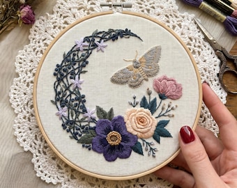 Op de afbeelding: Een ronde houten borduurring met een stoffen paneel voorzien van een handgestikt ontwerp van een maansikkel gevormd door donkerblauwe ranken en kleine lichtpaarse bloemen, een centrale mot en een cluster van paarse en perzikkleurige bloemen.