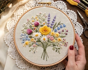 May include: A circular wooden embroidery hoop containing a fabric panel with a hand-stitched floral bouquet. The design features a central yellow flower surrounded by white daisies, pink clover, purple lavender, and blue bell-shaped flowers with green stems.