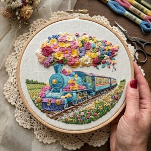 May include: A circular embroidery hoop containing a fabric panel with a detailed needlework design of a blue steam train travelling through a field. A large, colourful cloud of embroidered flowers in shades of pink, yellow, and blue rises from the train.