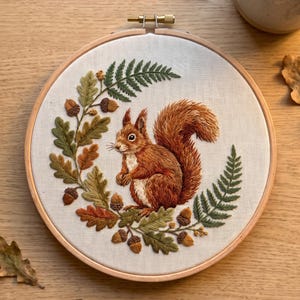 May include: A circular wooden embroidery hoop containing a fabric panel with a detailed thread-stitched design of a red squirrel surrounded by a wreath of green oak leaves, brown acorns, and fern fronds on a light cream-coloured background.