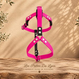 May include: A bright pink dog harness featuring black straps, silver-toned metal rivets, and black plastic buckles. The text Les Pattes Du Lynx HARNAIS CHIEN EN BIOTHANE is printed below the harness on the wooden surface.