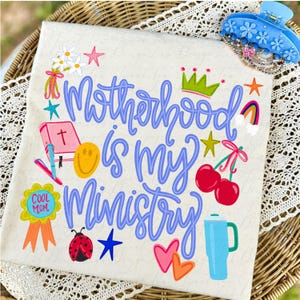 Può includere: Una maglietta color crema con la scritta "Motherhood is my Ministry" in blu. La maglietta è decorata con illustrazioni colorate tra cui una corona, ciliegie, un arcobaleno e una coccinella. Un fermaglio per capelli blu a forma di fiore si trova nell'angolo in alto a destra.