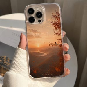 May include: Clear phone case featuring a scenic sunset landscape with a bird perched on a branch. The design includes warm orange and brown tones, with the text "CATMERA" at the bottom. The case is held by a person wearing a white sweater.