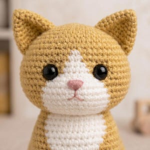 May include: A crochet cat plush toy featuring golden-yellow yarn with a white patch extending from the forehead down to the chest. The toy has two pointed ears, large black plastic safety eyes, and a small embroidered pink nose and mouth.