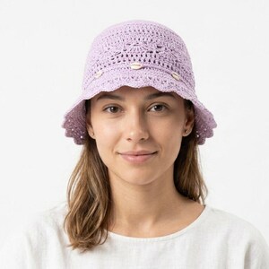 May include: A light lavender crochet bucket hat featuring an intricate openwork pattern and a scalloped brim. The hat is accented with a thin cord wrapped around the crown, decorated with three small, white, natural seashells spaced evenly.