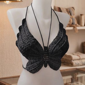 May include: A black crochet halter top shaped like a butterfly, featuring intricate textured stitching and thin braided straps that tie at the neck and back. The garment is displayed on a white mannequin, showcasing the detailed wing-like design.