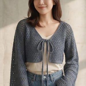 May include: A slate blue long-sleeve crochet bolero featuring an open-knit diamond pattern, a V-neckline, and a front tie closure. The garment has wide, flared sleeves and a cropped length, worn over a cream-colored camisole and blue denim jeans.