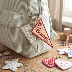 May include: Two triangular fabric pennant keychains with embroidered names Clark and Lennon attached to a striped backpack. Additional star and heart-shaped fabric keychains with embroidered letters S, E, and T are displayed nearby.
