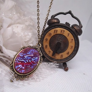 Puede incluir: Un collar de estilo vintage con un colgante ovalado rojo y azul y un pequeño reloj antiguo. El colgante tiene un marco y una cadena de color latón. El reloj es marrón con una esfera color crema y números negros.