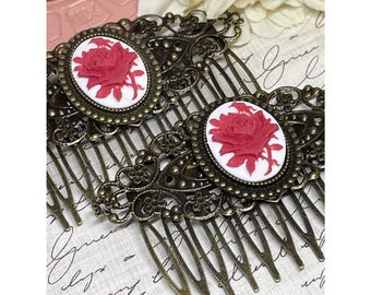 May include: Two antique bronze hair combs. Each comb features an oval cameo with a red rose design on a white background. The combs have intricate filigree detailing and are set against a backdrop of vintage script.
