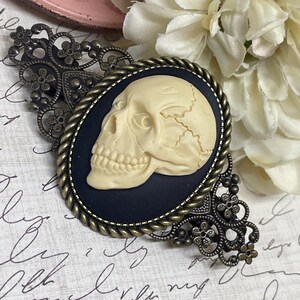 Könnte beinhalten: Ovale Cameo-Brosche mit einem cremefarbenen Totenkopf auf schwarzem Hintergrund, umrahmt von einem gedrehten Seildesign. Die Brosche ist in einer verzierten, bronzefarbenen Filigranarbeit gefasst.