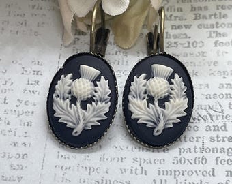 May include: A pair of oval-shaped earrings featuring a thistle design. The earrings have a dark blue background with a white thistle and leaf design. The earrings have a bronze-colored metal frame and lever-back closures.