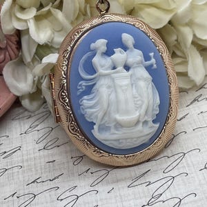 Op de afbeelding: Ovale goudkleurige medaillon met een lichtblauwe camee met twee witte figuren. De camee toont klassieke figuren met een vaas. De medaillon is open en toont scharnier en sluiting.