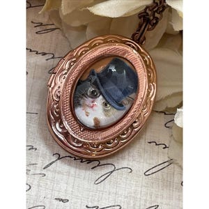 May include: A rose gold oval locket with a detailed design. The locket opens to reveal a glass cameo of a cat wearing a black top hat.