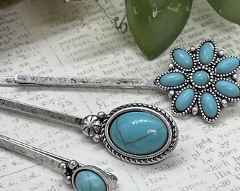 Op de afbeelding: Drie zilverkleurige haaraccessoires met turquoise gekleurde stenen. Eén is een haarspeld, een andere heeft een ovale steen en de derde heeft een bloemvorm. Op een vintage papieren achtergrond.