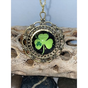 May include: A gold pendant necklace with a green four-leaf clover design in a glass cabochon. The pendant is surrounded by a decorative filigree frame.