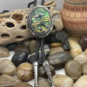 Op de afbeelding: Een bolo tie met een zilverkleurige ovale hanger met een inleg van groene, blauwe en gouden abalone-schelp. De stropdas is gemaakt van zwart gevlochten lederen koord met zilverkleurige uiteinden.