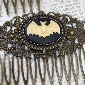 Op de afbeelding: Antieke bronzen haarspeld met een centraal ovale cameo met een cr&egrave;me vleermuis tegen een zwarte achtergrond. De haarspeld heeft ingewikkelde bloemen- en stipdetails, perfect voor Halloween of gotische stijlen.