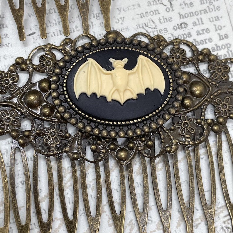 Op de afbeelding: Antieke bronzen haarspeld met een centraal ovale cameo met een cr&egrave;me vleermuis tegen een zwarte achtergrond. De haarspeld heeft ingewikkelde bloemen- en stipdetails, perfect voor Halloween of gotische stijlen.