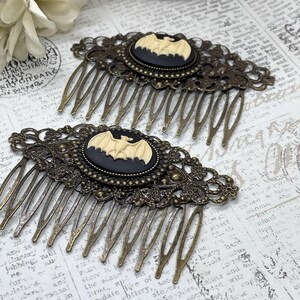Op de afbeelding: Twee antieke bronzen haar kammen. Elke kam heeft een zwarte ovale cameo met een cr&egrave;mekleurig vleermuisontwerp. De kammen hebben ingewikkelde filigraan details en lange, dunne tanden. Een gotisch of Halloween-thema accessoire.