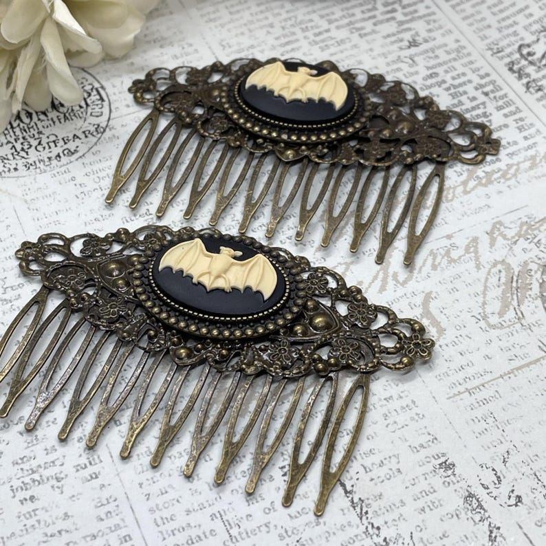 Op de afbeelding: Twee antieke bronzen haar kammen. Elke kam heeft een zwarte ovale cameo met een cr&egrave;mekleurig vleermuisontwerp. De kammen hebben ingewikkelde filigraan details en lange, dunne tanden. Een gotisch of Halloween-thema accessoire.