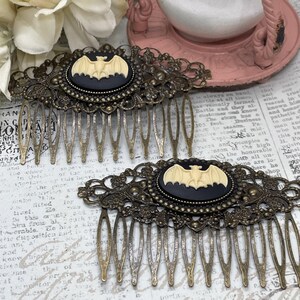 Op de afbeelding: Twee antieke bronzen haar kammen. Elke kam heeft een zwarte ovale cameo met een cr&egrave;mekleurig vleermuisontwerp. De kammen hebben ingewikkelde filigraan details en lange, slanke tanden.