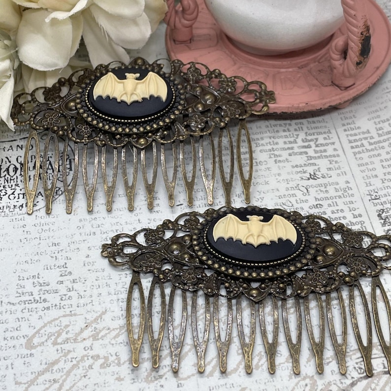 Op de afbeelding: Twee antieke bronzen haar kammen. Elke kam heeft een zwarte ovale cameo met een cr&egrave;mekleurig vleermuisontwerp. De kammen hebben ingewikkelde filigraan details en lange, slanke tanden.