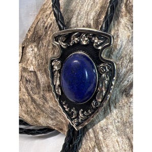 以下が含まれることがあります： 銀製のボロタイ。青い宝石が銀製の矢じり型のペンダントにセットされています。矢じりは銀のフィリグリーで装飾されており、黒い背景になっています。