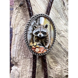 May include: A brown leather bolo tie with a silver oval pendant featuring a raccoon in a forest setting. The raccoon is looking directly at the viewer with a curious expression. The pendant is surrounded by a silver rope border.