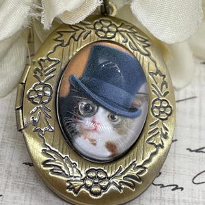 May include: Gold tone oval locket with a glass cameo of a cat wearing a top hat and monocle. The locket has a floral design around the edge.