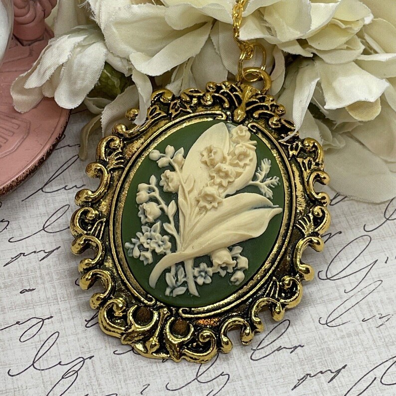 May include: A gold-tone pendant necklace features a cameo of a white lily of the valley flower on a green background. The cameo is set in an ornate oval frame.