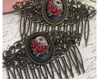 May include: Two antique bronze hair combs. Each comb features an oval cameo with a skull and red roses on a black background. The combs have intricate filigree detailing and long, thin teeth.