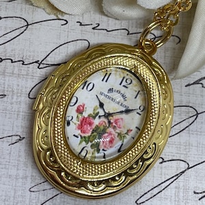 May include: Gold oval locket necklace with a floral clock face featuring a white background with pink roses and the text "Maraville Senteurs de Rose".