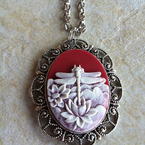 May include: A silver pendant necklace with a red cameo featuring a white dragonfly and flowers. The cameo is surrounded by a silver filigree frame.