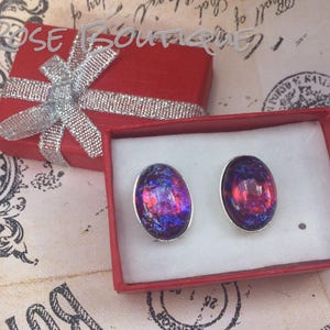 May include: A pair of oval-shaped stud earrings with a silver border, presented in a red gift box. The earrings feature a vibrant mix of red and blue colors. The gift box is tied with a silver ribbon.