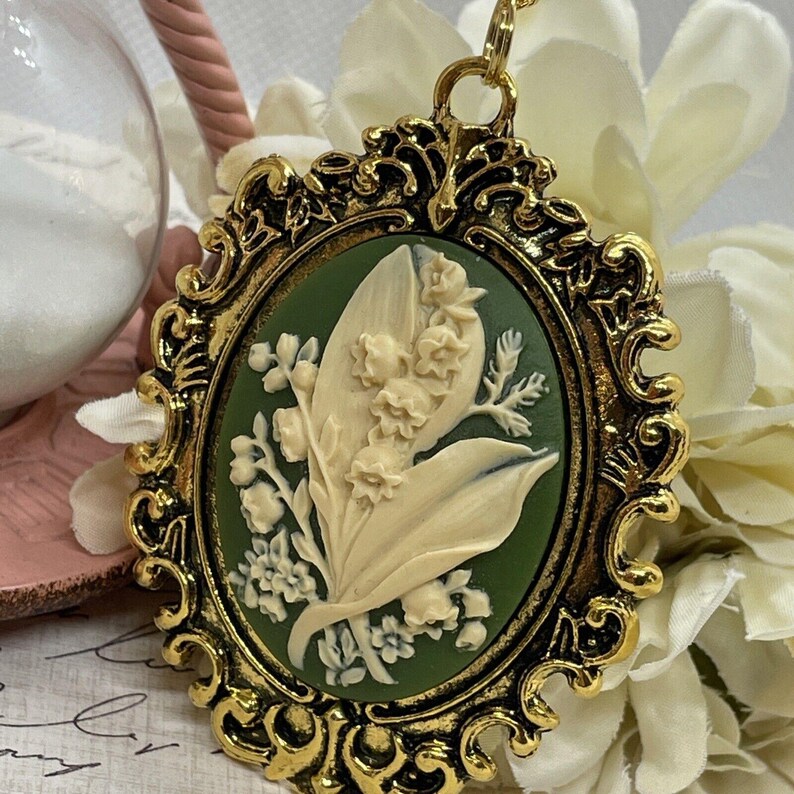 May include: A gold-tone pendant with a cameo of a lily of the valley flower in white on a green background.
