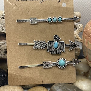 Puede incluir: Conjunto de tres pinzas para el pelo plateadas con piedras turquesa. Cada pinza presenta un diseño de flecha con un acento de piedra turquesa diferente. La pinza del medio tiene un diseño de pájaro.