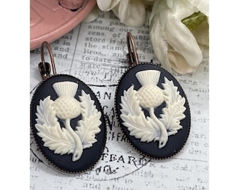 May include: A pair of oval-shaped earrings with a black background and a cream-colored thistle design. The earrings have a bronze-colored hook closure. The thistle is surrounded by leaves.