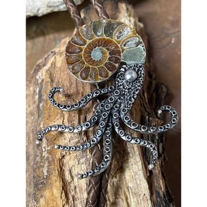 Op de afbeelding: Een bolo tie met een zilveren octopus met een gefossiliseerde ammoniet-schelp. De octopus heeft een getextureerd lichaam en gekrulde tentakels. De ammoniet-schelp is bruin en heeft een spiraalvormig patroon.