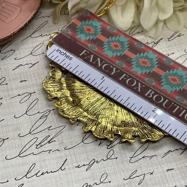 May include: A gold-toned metal pendant with a scalloped edge. The pendant is being measured with a ruler that reads "inches" and has the text "Fancy Fox Boutique" printed on it.